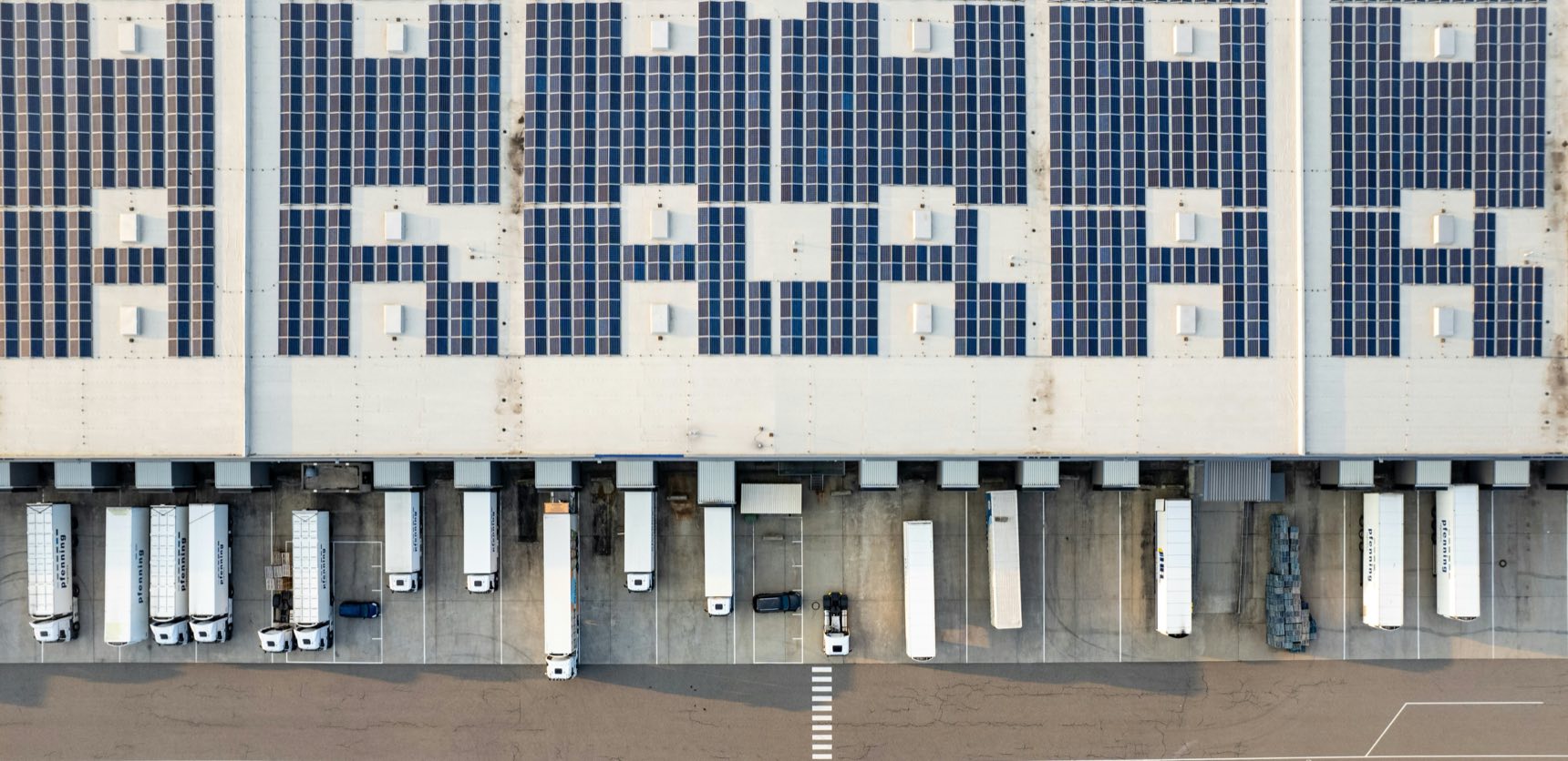 An aerial view of a distribution centre. Solar panels cover the roof. Articulated trailers stand in loading bays along its length, with several bays empty.